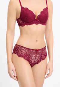 Reggiseno in pizzo bordeaux con motivi floreali e dettaglio a fiocco al centro; abbinato a slip in pizzo coordinati con disegni elaborati.