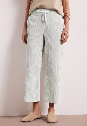 Personne portant un pantalon beige clair rayé avec un cordon et des chaussures beiges à enfiler, debout contre un mur clair uni.