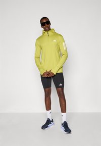 Limonengrünes langärmliges Sportshirt mit Kapuze und Reißverschluss, kombiniert mit schwarzen Shorts, weißen Akzenten und dunkelblauen Laufschuhen mit weißen Details.