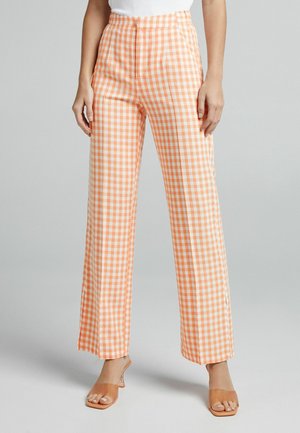 Oranje en witte ruitpatroon broek met hoge taille, wijde pijpen en een soepele textuur; gecombineerd met doorzichtige hielsandalen.