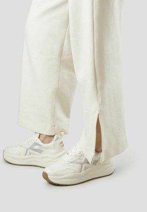 Sneakers bianche con suole spesse indossate con pantaloni crema a gamba larga con spacchi laterali, mostrati su uno sfondo grigio chiaro.