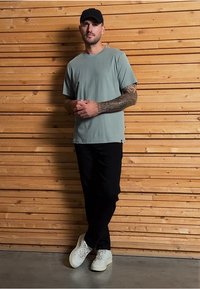 Světle modré tričko s krátkým rukávem, černé kalhoty, bílé tenisky, stojící proti dřevěné zdi. Tetování viditelné na rukou.