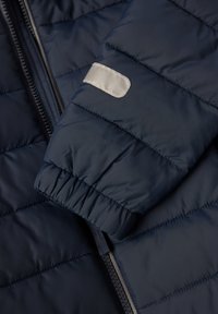 Marineblå puffjakke med kviltet design. Har elastiske manchetter, et sølvreflekterende mærke og en frontlynlås. Glat tekstur.