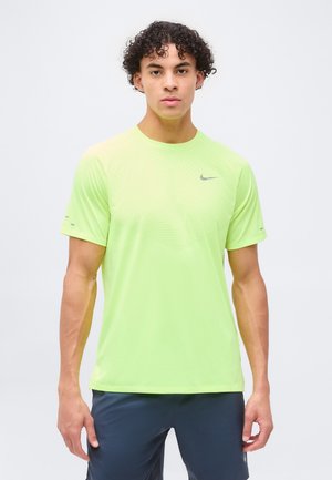 Junger Mann mit lockigem Haar, der ein neongelbes Nike-Sportshirt und dunkelblaue Shorts trägt, vor einem schlichten hellen Hintergrund.