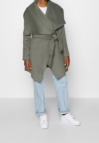 Cappotto avvolgente verde oliva con collo drappeggiato, cintura a cravatta e maniche lunghe, abbinato a jeans azzurri chiari e sneakers bianche. Texture liscia.