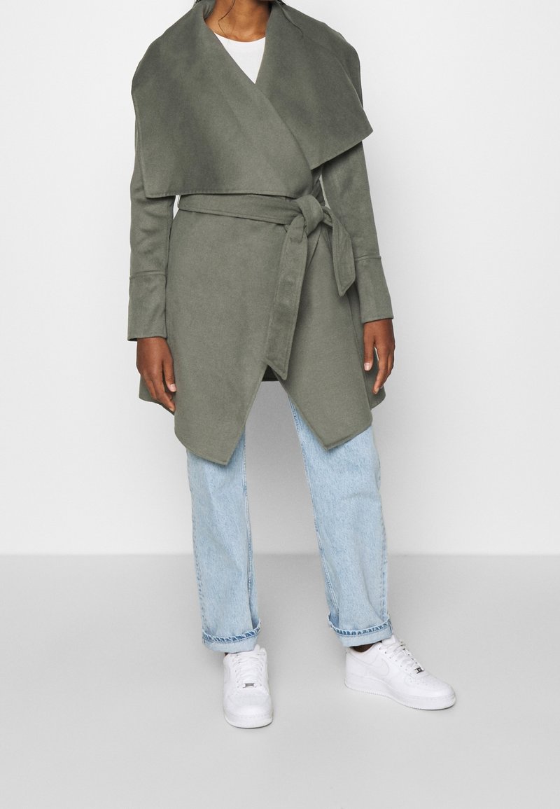 Cappotto avvolgente verde oliva con collo drappeggiato, cintura a cravatta e maniche lunghe, abbinato a jeans azzurri chiari e sneakers bianche. Texture liscia.