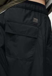 Μαύροι παντελόνια εργασίας με ελαστική μέση και μεγάλη τσέπη με καπάκι που φέρει ετικέτα της μάρκας TAION στο ισχίο.