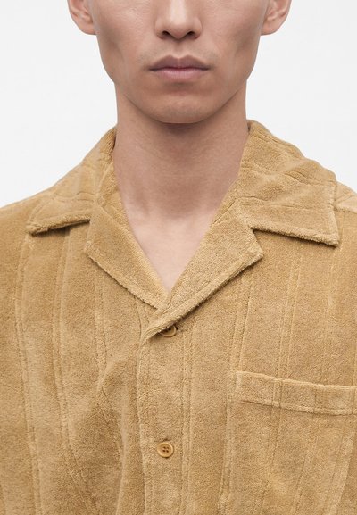 Gros plan d'une personne portant une chemise texturée marron clair avec un col, une patte de boutonnage à l'avant et une poche poitrine, sur un fond blanc uni.