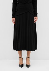 Jupe plissée noire avec une ceinture structurée comportant des poches, un tissu fluide et une longueur mi-mollet. Portée avec des chaussures à talons hauts noires.