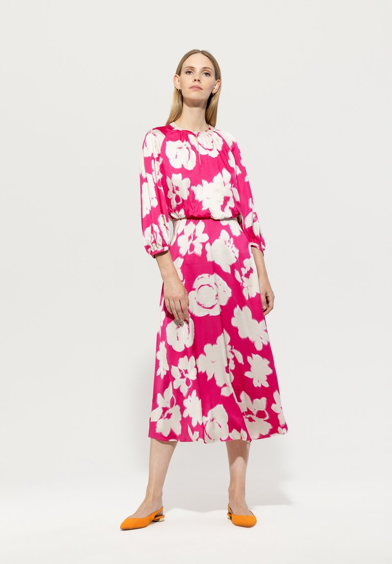 Blumenkleid in kräftigem Pink mit großen weißen Blumen, locker geschnitten, mit langen Ärmeln und einem fließenden Rock. Kombiniert mit orangen Ballerinas.