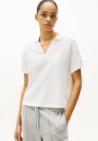 Witte polo met korte mouwen en een v-hals, in een relaxte pasvorm. Zacht materiaal, met een kleine logo-accent op de manchet. Gecombineerd met grijze joggingbroeken.