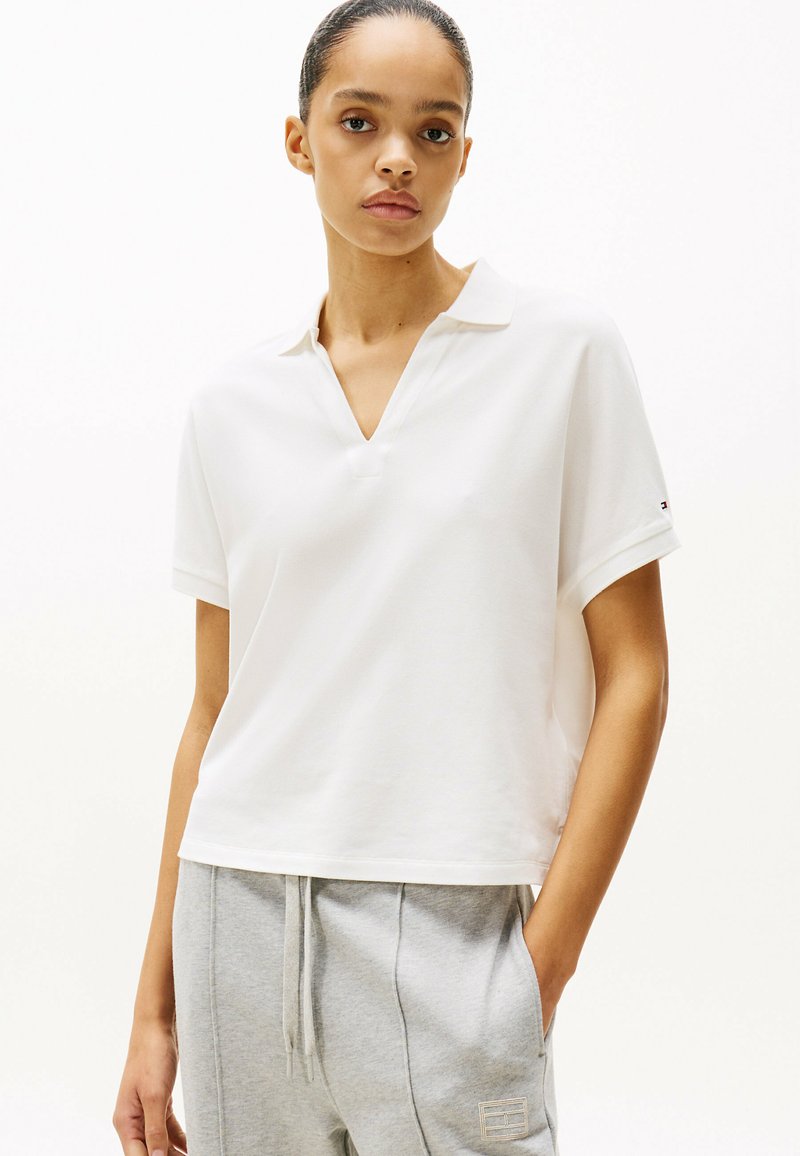 Witte polo met korte mouwen en een v-hals, in een relaxte pasvorm. Zacht materiaal, met een kleine logo-accent op de manchet. Gecombineerd met grijze joggingbroeken.