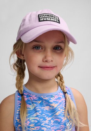Lila Baumwollmütze mit einem schwarzen rechteckigen Logopatch, getragen zu einem ärmellosen Oberteil in Blau und Pastellmustern. Kind mit blonden Haaren in Zöpfen.
