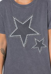 Grå T-shirt med to stjerne-applikationer, en stor og en lille, med sølvnitter der omkranser hver stjerne og et blødt, struktureret stof.