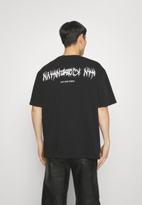 Černé bavlněné tričko s výraznými bílými grafickými nápisy na zadní straně. Volný střih s krátkými rukávy a kulatým výstřihem.