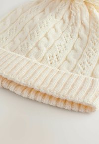 Crème-kleurige gebreide beanie met kabel- en ribbelpatronen. Slouchy ontwerp met een zachte, textuur die je kunt voelen. De manchetten zijn iets omgeslagen.