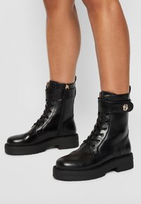 Bottes de combat en cuir noir avec devant lacé, ferrures dorées, et détail de sangle à la cheville. Dotées d'une semelle épaisse et d'une fermeture éclair latérale.