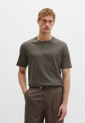 Jonge man met lichtbruin haar, gekleed in een effen olijfgroen T-shirt met korte mouwen en bruine broek met trekkoord, staand met één hand in zijn zak.