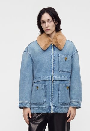 Ung kvinde iført en lyseblå denimjakke med en brun pelskrave og guldknappede lommer mod en ensartet hvid baggrund.