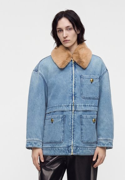 Jeune femme portant une veste en jean bleu clair avec un col en fourrure beige et des poches à boutons dorés sur un fond blanc uni.