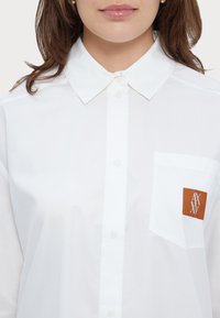 Camisa blanca de botones con cuello puntiagudo y un bolsillo en el pecho izquierdo que presenta un parche de cuero marrón con un logo. Tejido suave y ligero.
