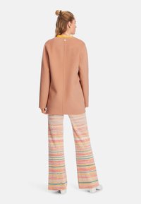 Beiger Mantel mit sauberem Design, der einen runden Ausschnitt und eine Knopfdetail auf der Rückseite aufweist. Kombiniert mit bunten, im Zickzackmuster gestalteten weit geschnittenen Hosen.