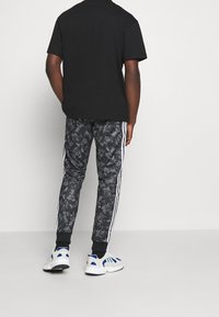 Person som bär svarta grafiska joggarbyxor med vita sidostråk, vita sneakers med blå accenter och en enkel svart T-shirt med kort ärm, står.