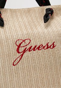Sac en tissu beige tissé avec des poignées noires à motifs et un logo "Guess" brodé en rouge en écriture cursive.