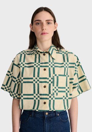 Femme aux cheveux bruns lisses porte une chemise beige à manches courtes avec un motif à carreaux verts et un jean bleu foncé sur un fond uni.