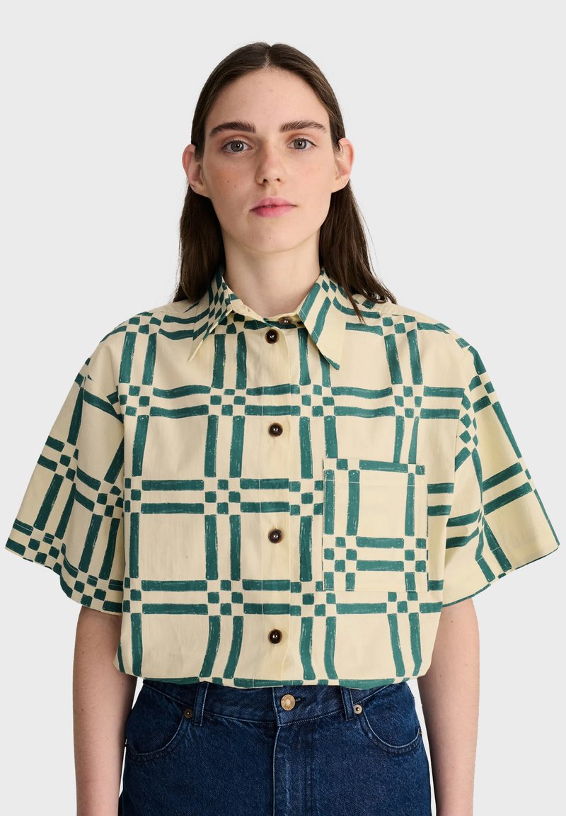 Femme aux cheveux bruns lisses porte une chemise beige à manches courtes avec un motif à carreaux verts et un jean bleu foncé sur un fond uni.