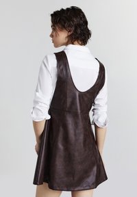 Donna con capelli corti scuri, che indossa una camicia bianca e un vestito in pelle marrone scuro senza maniche, in piedi davanti a uno sfondo chiaro e semplice.