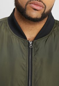 Giacca bomber verde oliva con colletto a costine nero e chiusura con zip. Presenta una texture liscia e un leggero luccichio.