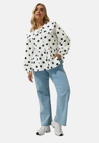 Blusa bianca con grandi pois neri, colletto a volant e vita cinchata, abbinata a jeans a gamba larga blu chiaro e sneaker bianche.