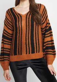 Frau trägt einen lockeren orangen Strickpullover mit vertikalen und horizontalen schwarzen Streifen, kombiniert mit schwarzen Samenhosen.