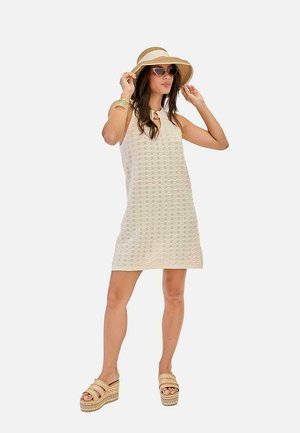 Mujer con vestido sin mangas beige con estampado, sombrero de ala ancha, gafas de sol, pulsera y sandalias de plataforma posando con las manos ajustando el sombrero.