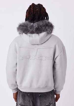 Persoon met donker, gedraaid haar draagt een lichtgrijze hoodie met bont afgewerkte capuchon en "PXP" in reliëf op de achterkant, gecombineerd met een donkere spijkerbroek.
