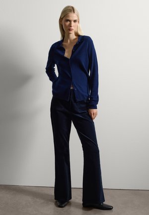 Mujer de pie contra una pared blanca, con un cárdigan azul marino, pantalones oscuros de pierna ancha y zapatos negros, con una mano en el bolsillo.