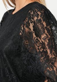 Czarna koronkowa bluzka z kwiatowym wzorem, przezroczystymi długimi rękawami i jednolitą czarną częścią. Teksturowany, lekki materiał z okrągłym dekoltem.