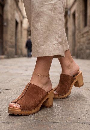 Piedi che indossano zoccoli con tacco aperto in suede marrone con suola in legno e tacco a blocco, pantaloni beige corti e un braccialetto alla caviglia d'oro su una strada lastricata.