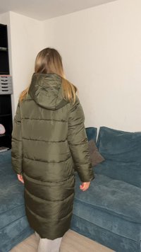 Ilgas, pūstas alyvuogių žalsvos spalvos paltas su gobtuvu, su horizontaliomis siūlėmis, matomas iš nugaros. Minkšta tekstūra, siekianti vidurį blauzdos.