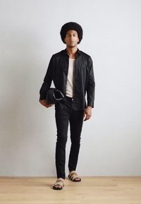 Mustapaita, valkoiset raidat paidan alla, mustat slim-fit-housut, vaaleat sandaalit, pitämässä harmaata esinettä, seisomassa puulattialla.