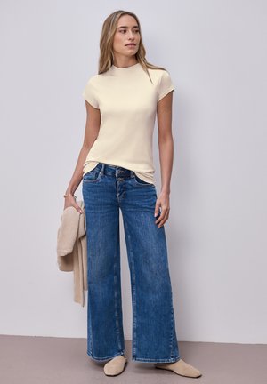 Nainen pukeutuneena kermaiseen lyhythihaiseen toppiin ja leveälahkeisiin sinisiin farkkuihin, kädessä beige neule, seisoo vaalean ja yksinkertaisen seinän edessä.