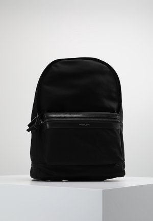 Schwarzer Stoffrucksack mit vorderer Reißverschlusstasche und kleinem "Michael Kors Est. 1981"-Logo, auf einem weißen Podest vor einem einfarbigen Hintergrund platziert.