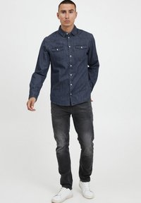 Camisa de mezclilla oscura con mangas largas, cierre de botones en la parte delantera, dos bolsillos en el pecho y un dobladillo ligeramente curvado. Llevada con jeans grises y zapatillas blancas.