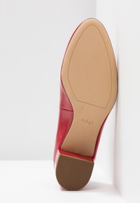Chaussure en cuir verni rouge montrant une surface lisse et brillante avec un bout effilé. La semelle est beige avec une texture antidérapante et un logo.
