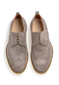 Graue Wildlederschuhe mit einem Wingtipp-Design, das dekorative Perforationen, flache Schnürsenkel und eine kontrastierende Gummisohle aufweist. Der hintere Tab hat einen gestreiften Akzent.