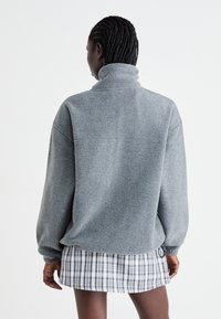 Grauer Fleece-Pullover mit hohem Kragen, lässiger Passform und hängenden Schultern, kombiniert mit einem karierten Minirock in Schwarz und Weiß.