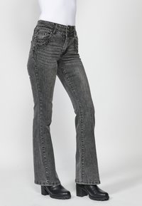 Pantalones vaqueros negros acampanados con un acabado texturizado, que presentan costuras decorativas y una cintura alta. Combinados con botas negras de tacón grueso.