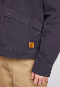 Dark grey jacket with a textured finish, featuring a large front pocket and a prominent orange Timberland logo patch.