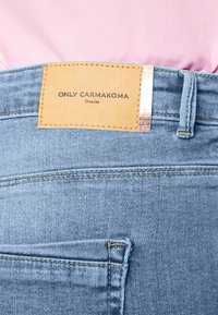 Šviesiai mėlynos džinsinės kelnės su šviesiai rudos odos etiketėmis, jų paviršiuje įspausti žodžiai "ONLY CARMAKOMA" ir "Denim", su subtiliais siuvinėjimais ir užpakaline kišenėle.